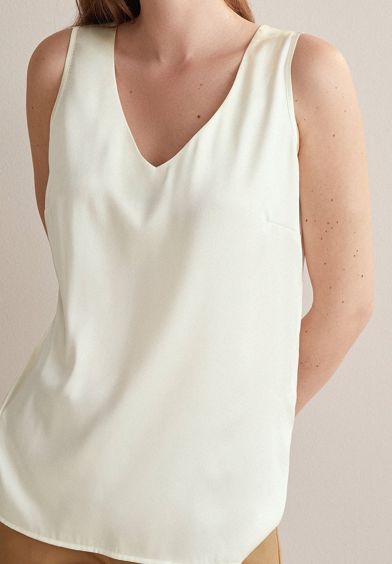 Top senza maniche a scollo a V in tessuto crema chiaro. Texture liscia con una vestibilità rilassata e un piccolo dettaglio tasca sul lato sinistro.