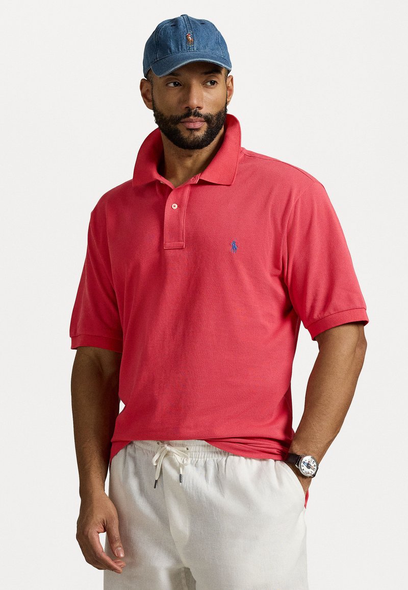 Rde polo majica z modrim vezenim logotipom, v kombinaciji z belimi hlačami na vrvico in svetlo modro denim kapo. Ure z black pasom.