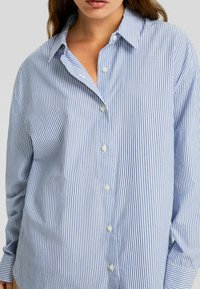 Kvinna som bär en lös, långärmad skjorta med blå och vita vertikala ränder, där den översta knappen är öppen.