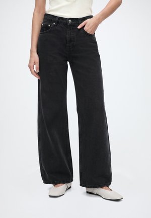 Personne portant un jean noir taille haute à jambes larges et des chaussures plates blanches, avec une main dans une poche avant, sur un fond clair uni.