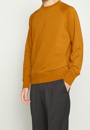 Mann trägt ein langärmeliges Senfgelbes Sweatshirt und dunkelgraue Hose, mit einer Hand in der Hosentasche, vor einem einfarbigen hellen Hintergrund.