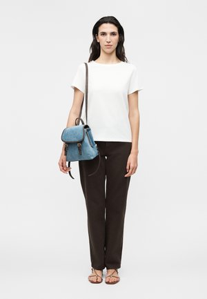 Naine seisab näoga ette, seljas valge lühikeste varrukatega särk, mustad püksid, pruunid sandaalid ning ühe õla peal sinine seljakott.