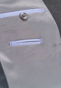 Grauer Stoff mit einem hellblauen Akzent, der über einen Knopf und eine kleine Tasche mit blauen Nähdetails verfügt. Glatte Textur, maßgeschneidertes Design.