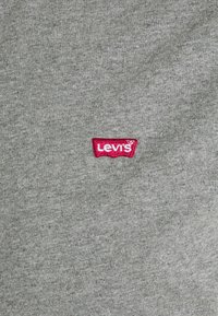 Tissu gris avec un petit écusson rouge et blanc Levi’s cousu près du centre. Aucun autre objet ou motif visible.