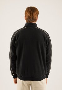 Černá fleecová bunda s vysokým límcem, s bílým lemováním podél ramen a elastickými manžetami. Měkká textura, volný střih.