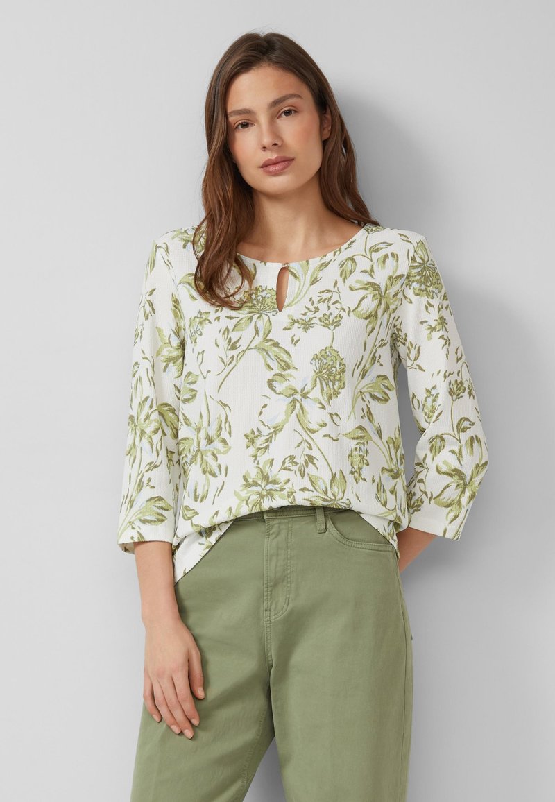 Kobieta ubrana w białą bluzkę z długim rękawem z zielonym nadrukiem kwiatowym oraz jasnooliwkowe spodnie, stojąca na tle jednolitego jasnoszarego tła.