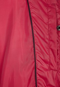 Interior de chaqueta de nylon roja con textura suave, costuras horizontales, ribete negro en el zipper y un bolsillo lateral.