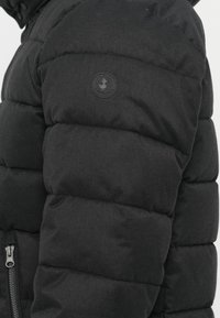 Svart puffjacka med kviltad struktur, med en zippered ficka och rund logotyp patch på axeln. Hållbart material, isolerad design.