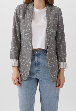 Kobieta nosząca szarą kratowaną marynarkę z podwiniętymi rękawami, biały krótki top i jasnoniebieskie jeansy z wysokim stanem na jednolitym tle.