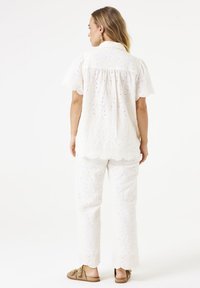 Witte blouse en broekset met bloemenborduurwerk en gegolfde randen. De stof is textuur, met kleine uitsparingen en subtiele gouden accenten.