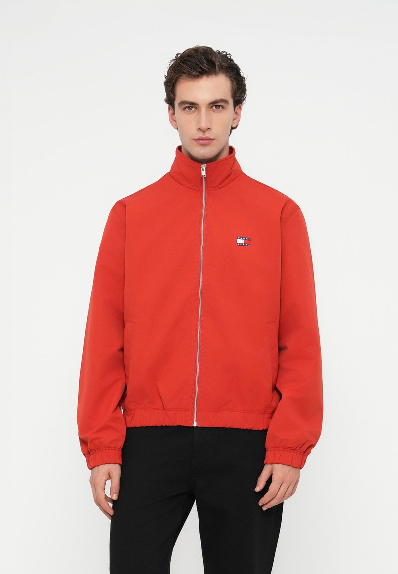 Κόκκινη bomber τζάκετ με στέρεο γιακά, μπροστινό φερμουάρ, ελαστικές μανσέτες και τελείωμα, με μικρό μπλε και λευκό λογότυπο στο στήθος.