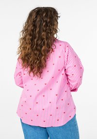 Lyserød stribet bluse med små røde hjerter, lange puffede ærmer og en buet kant. Kombineret med lyseblå denimjeans.