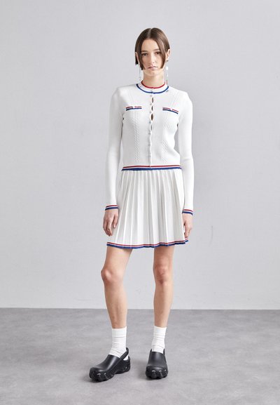 Robe tricotée blanche à manches longues, ornée de rayures rouges et bleues, jupe plissée et accents contrastants. Des chaussures noires complètent le look.