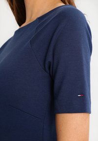 Närbild av en person som bär en mörkblå kortärmad skjorta med en liten röd, vit och blå broderad logotyp på ärmen.