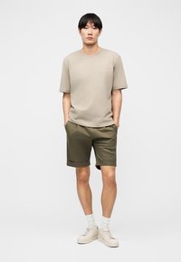 Νεαρός άντρας στέκεται με τα χέρια στις τσέπες, φορώντας μπεζ μπλούζα, λαδί σορτς, λευκές κάλτσες και μπεζ αθλητικά παπούτσια, σε απλό φόντο.