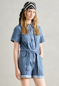 Denim jumpsuit med korte ermer, knapp foran, to brystlommer, belte i midjen og rullede mansjetter i lyseblå vask. Hodeplagg med svart og hvit stripet mønster.