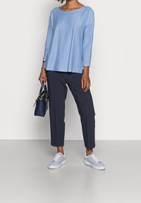 Jasnoniebieska bluzka w paski z długim rękawem, połączona z ciemnymi spodniami i jasnoniebieskimi trampkami, z małą ciemną torebką trzymaną w ręku.