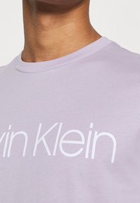 Camiseta de algodón color lila claro con cuello redondo y un gran logo blanco de "Calvin Klein" en el frente. Textura suave y diseño sencillo.