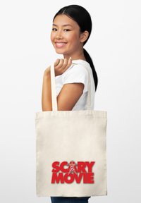 Uśmiechnięta kobieta z czarnymi włosami trzyma beżową torbę z napisanym na niej czerwonym tekstem "Scary Movie" z stylizowaną literą A na białym tle.