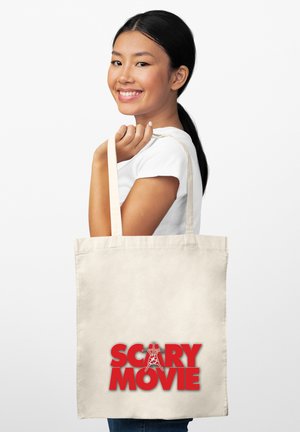Uśmiechnięta kobieta z czarnymi włosami trzyma beżową torbę z napisanym na niej czerwonym tekstem "Scary Movie" z stylizowaną literą A na białym tle.