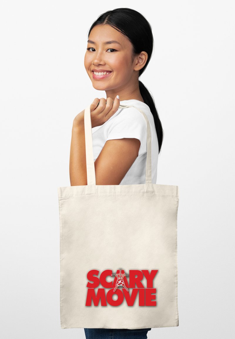 Uśmiechnięta kobieta z czarnymi włosami trzyma beżową torbę z napisanym na niej czerwonym tekstem "Scary Movie" z stylizowaną literą A na białym tle.