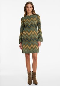 Woman wearing a knee-length zigzag patterned dress in earthy tones with long sleeves and brown studded ankle boots, standing on white background.