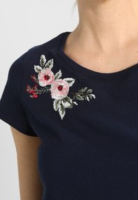 Primer plano de una camiseta azul marino con flores bordadas en rosa y blanco y hojas verdes cerca del escote del hombro izquierdo.