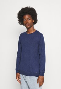 Navy blue long-sleeve sweater made from textured fabric, featuring a crew neckline and ribbed cuffs, paired with light denim jeans.