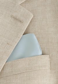 Chaqueta de traje beige texturizada con un pañuelo de bolsillo azul claro visible en el bolsillo del pecho. Diseño ajustado con un detalle de costura simple.