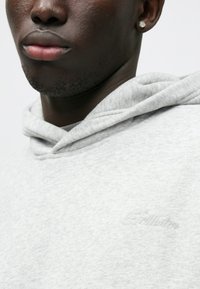 Szara bluza z kapturem o miękkiej fakturze, z subtelnym haftowanym logo w tym samym kolorze na piersi. Widoczny detal ściągacza na kapturze.