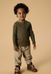 Olivengroen geribbelde longsleeve shirt, beige en bruine geruite broek, bruine veterschoenen, staand tegen een neutrale achtergrond.