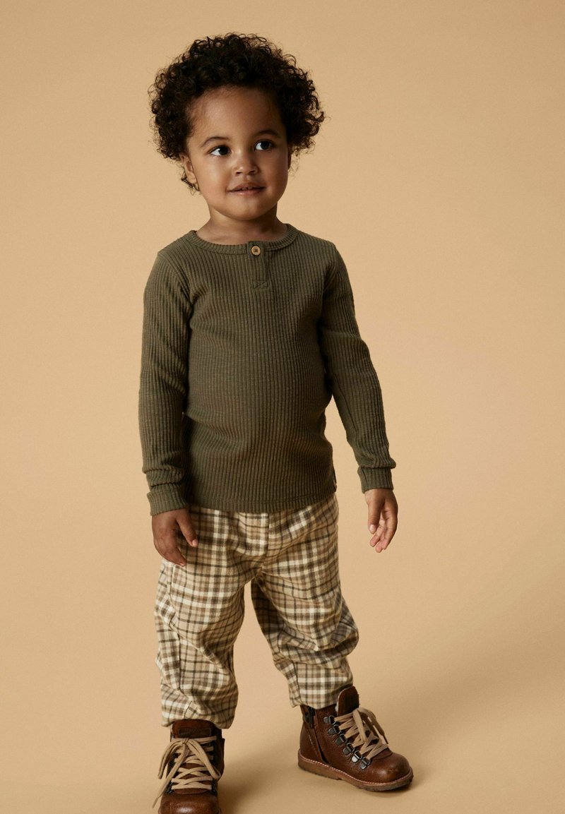 Olivengroen geribbelde longsleeve shirt, beige en bruine geruite broek, bruine veterschoenen, staand tegen een neutrale achtergrond.