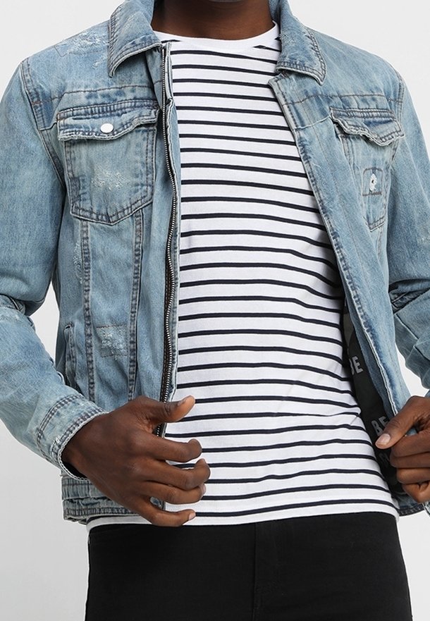 Person trägt eine hellblaue Jeansjacke über einem weiß-schwarz horizontal gestreiften Shirt und richtet die Jacke mit beiden Händen.