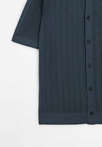 Cardigan en maille bleu foncé à manches courtes, texture côtelée verticale et boutons noirs sur le devant.