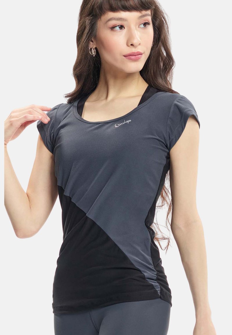 Donna che indossa una maglietta sportiva aderente a maniche corte bicolore grigio e nero con un piccolo logo sul petto, che guarda di lato.