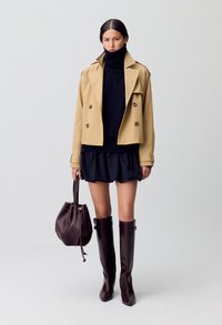Manteau trench beige à double boutonnage, pull col roulé bleu marine, mini-jupe évasée foncée, bottes hautes bordeaux et sac seau assorti.