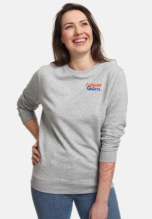 Frau lächelt, trägt einen grauen Pullover mit dem Text "Florida Gators" auf der Brust und blaue Jeans, steht mit einer Hand in der Hüfte.
