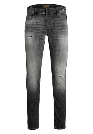 Person iført sort sweatshirt med kranie- og skateboardgrafik, falmede revnede grå jeans og sorte og hvide sneakers, der læner sig sidelæns.