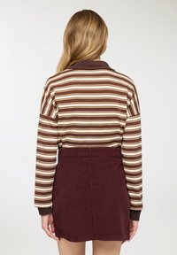 Donna con capelli biondi lunghi che indossa una maglia a maniche lunghe a righe marroni e crema e una gonna a vita alta marrone scura, vista di schiena.