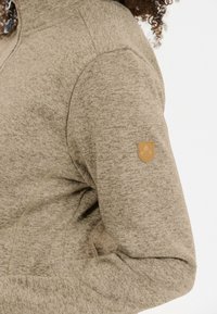 Khaki fleecejacka med en texturerad yta, halvdragkedja och en brun logotyp på vänster axel.