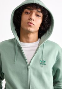 Camisola com fecho éclair em verde menta, com capuz ajustável, logotipo verde bordado e textura de tecido macio, usada sobre uma camisa branca.