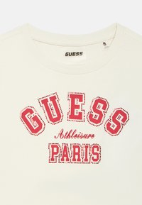 Λευκό T-shirt με κόκκινο τυπωμένο κείμενο «GUESS Athleisure PARIS» με διακοσμητικές τελείες, στρογγυλή λαιμόκοψη και ετικέτα με μέγεθος 8.