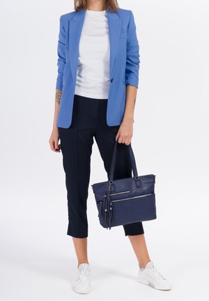 Žena v modrém saku, bílé košili, černých krátkých kalhotách a bílých teniskách držící tmavě modrou kabelku za rukojeti.