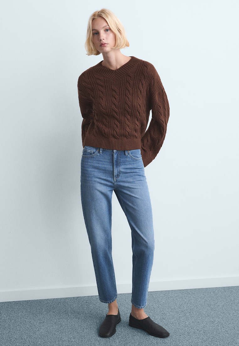 Jeune personne aux cheveux blonds courts, debout, portant un pull en maille torsadée marron, un jean bleu taille haute et des chaussures noires sans lacets.