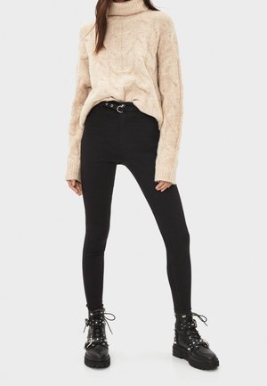 Suéter de cuello alto beige tejido con cable, ajuste relajado; jeans pitillo negros de cintura alta; botas negras gruesas estilo militar con tachuelas plateadas.