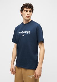 Νέος άνδρας με σγουρά μαλλιά που φορά μπλε ναυτικό μπλουζάκι New Balance Athletics και καφέ κοντά παντελόνια, κοιτάζοντας προς τα αριστερά μπροστά σε απλό φόντο.