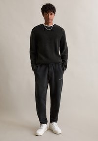 Schwarzer Ripp-Pullover mit langen Ärmeln, kombiniert mit schwarzen Sweatpants mit Kordelzug und subtil geprägtem Logo. Weiße Sneakers runden das Outfit ab.