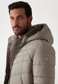 Jachetă puffer de culoare bej deschis, cu glugă, având detalii cusute, închidere cu fermoar și un logo triunghiular verde pe umărul stâng.