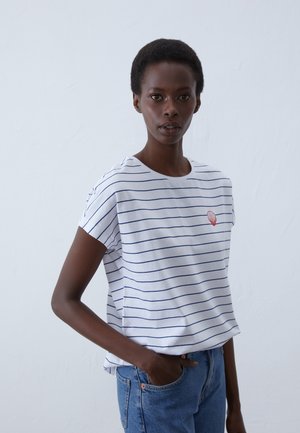 T-shirt en coton blanc avec des rayures navy, manches courtes, et une broderie en forme de coquillage rouge sur la poitrine, associé à un jean bleu.