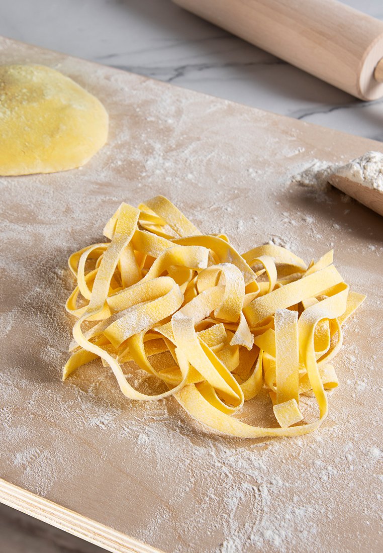 Vers gemaakte fettuccine pasta in een kluwen, bestoven met bloem, op een houten oppervlak met een deegroller en deeg op de achtergrond.